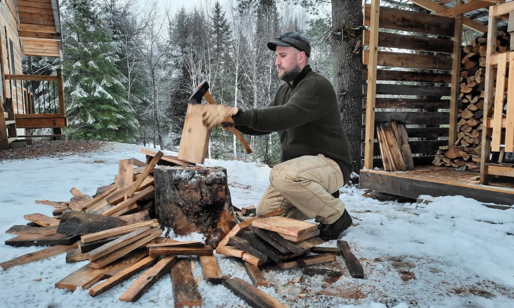 Chris using axe to chop firewood into a pile wearing green woolpower full zip jacket