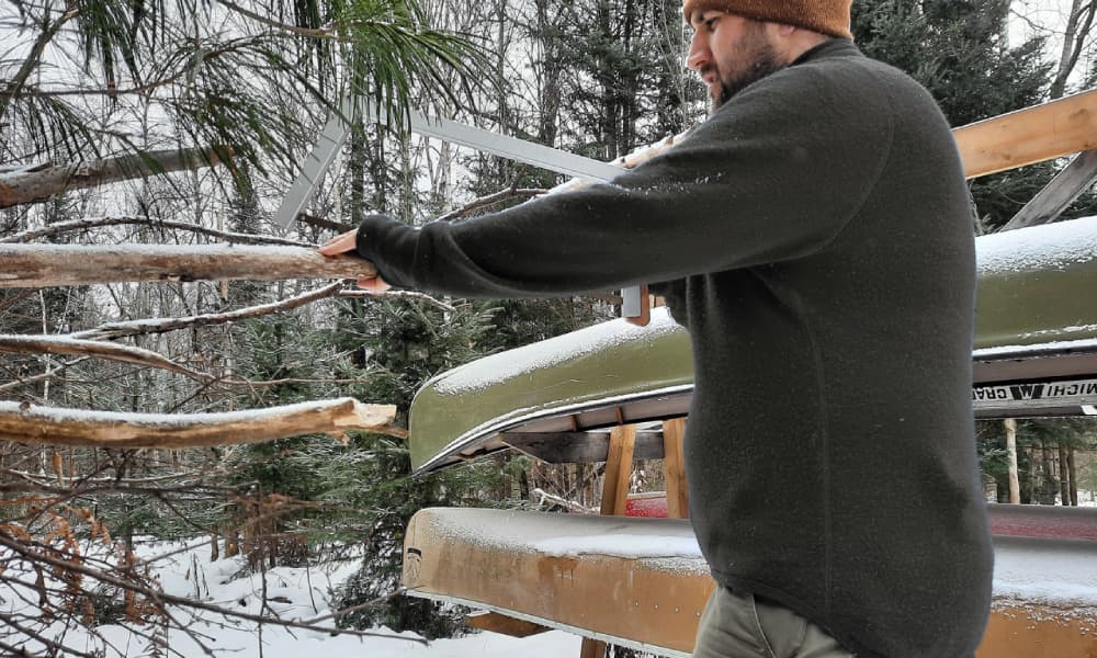 Chris cutting wood in green Woolpower full zip jacket with canoes in background